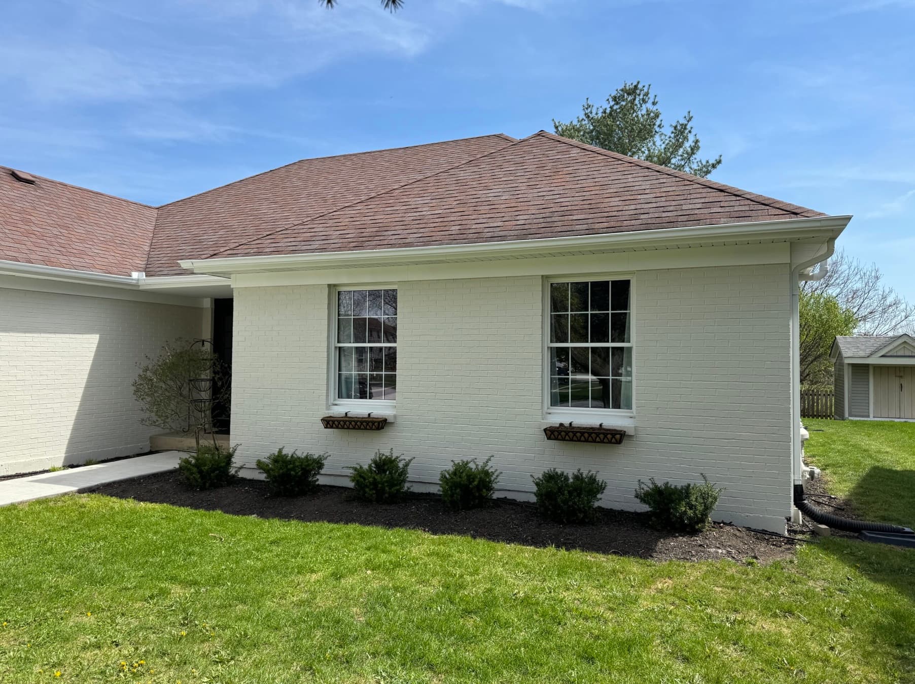 White brick house with brown roof, two windows with window boxes, and green bushes.